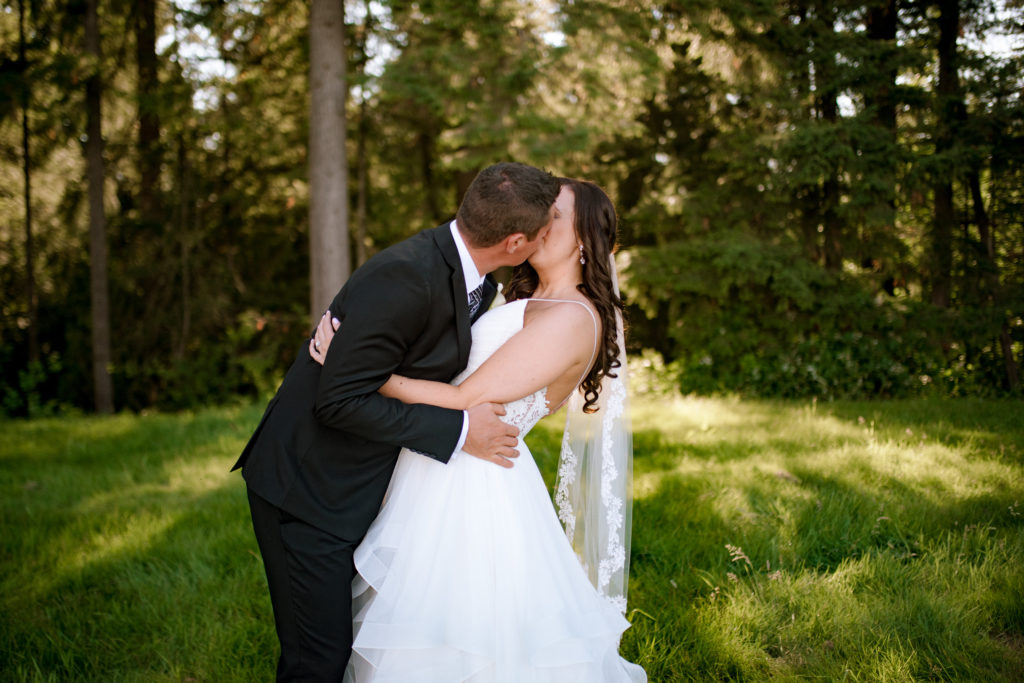 A newly married Bride and Groom embrace and kiss with lush green grass and woods in the background
