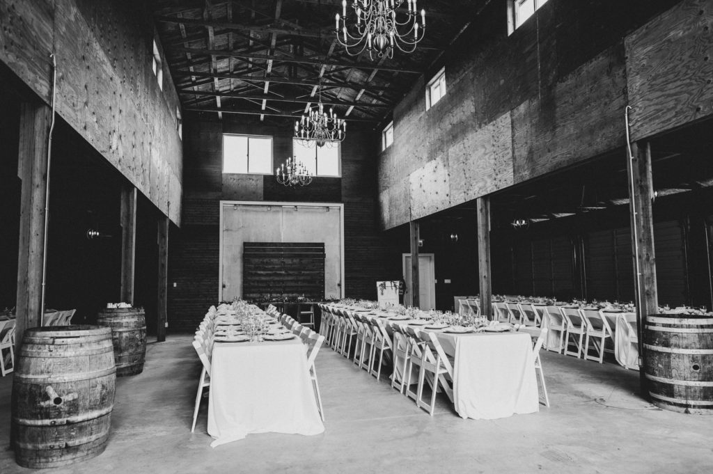 A black and white image of two long farm tables and white chairs set for a wedding reception among wine barrels and chandeliers inside the barn at Hopcott Farms