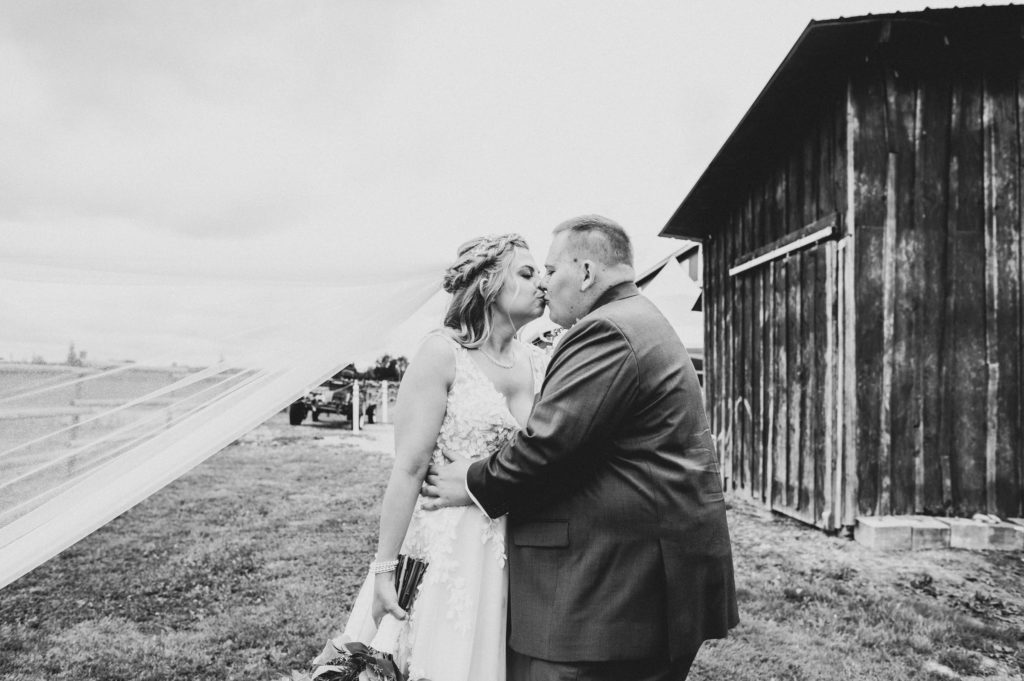 A bride and groom kiss by a rustic barn and long veil flowing to the side at Hopcott Farms