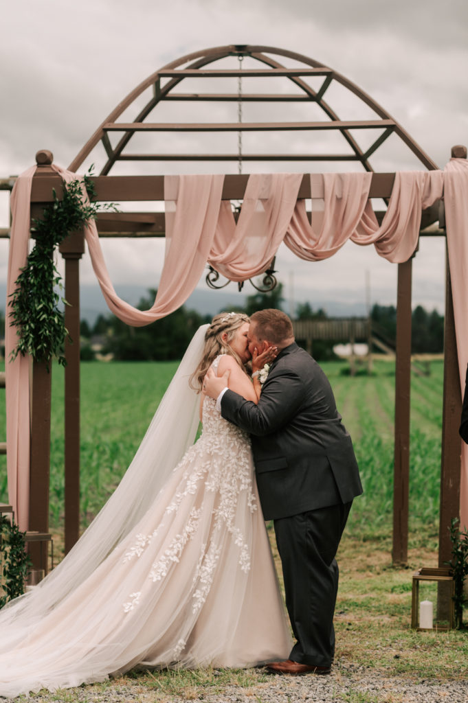 A bride and groom kiss underneath a simple wooden pergola with blush pink fabric, greenery and a country chandelier draped around them.