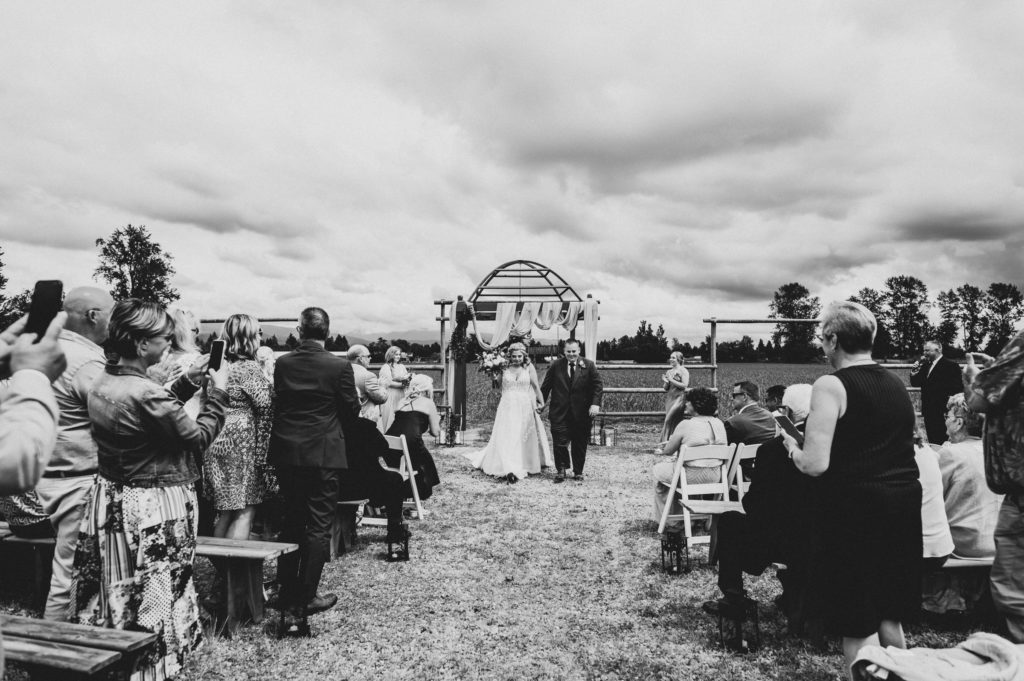 A bride and groom walk away from the wooden arches and towards their wedding ceremony guests at Hopcott Farms under a cloudy sky.