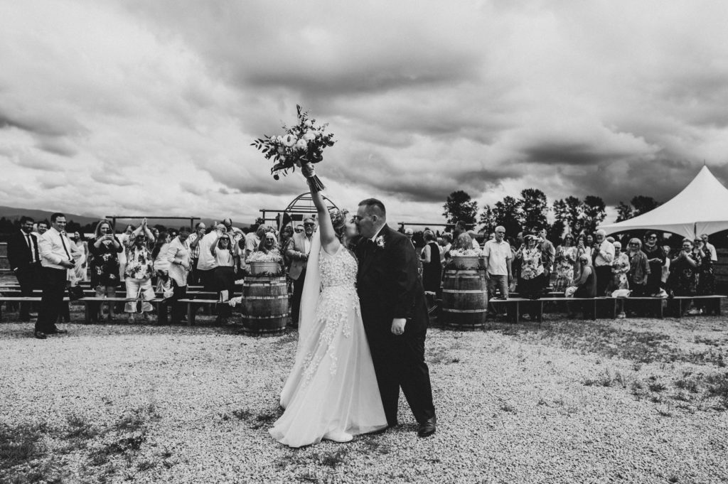 A newly married bride and groom kiss with the bridal bouquet in the air in front of the open air farm ceremony space on a cloudy day