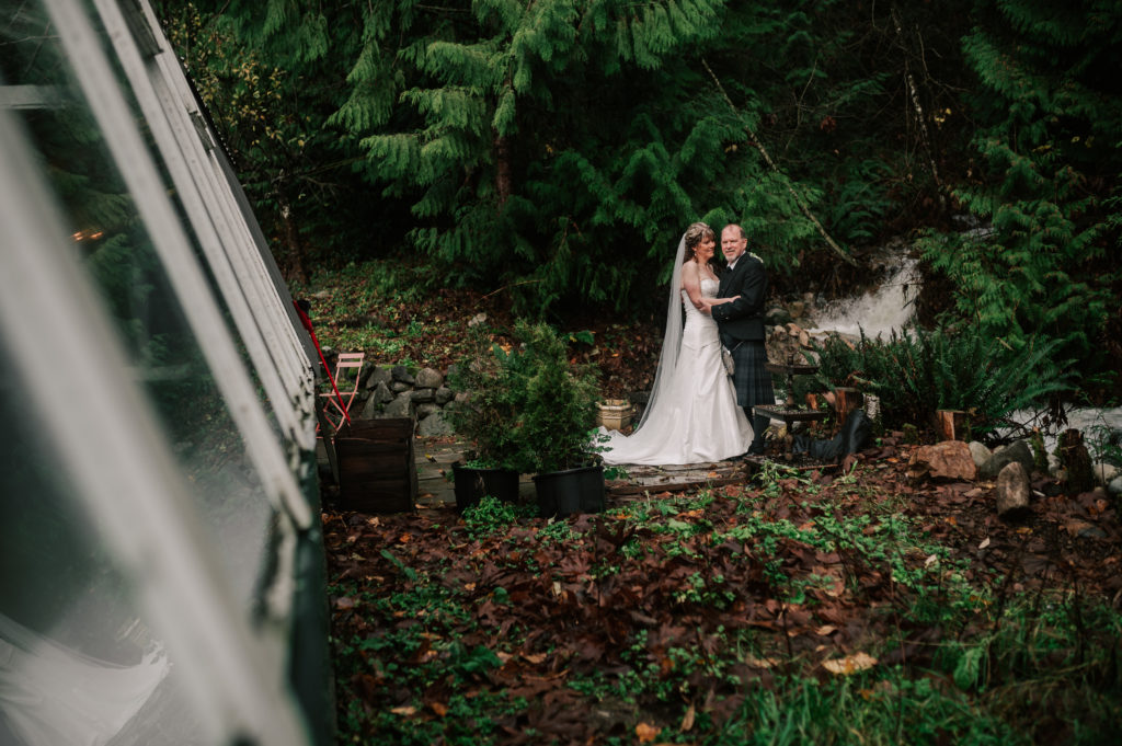 Alternative Vancouver wedding venue The Greenhouse view of outside the venue with bride and groom standing together near a waterfall and stream among green cedars and ferns